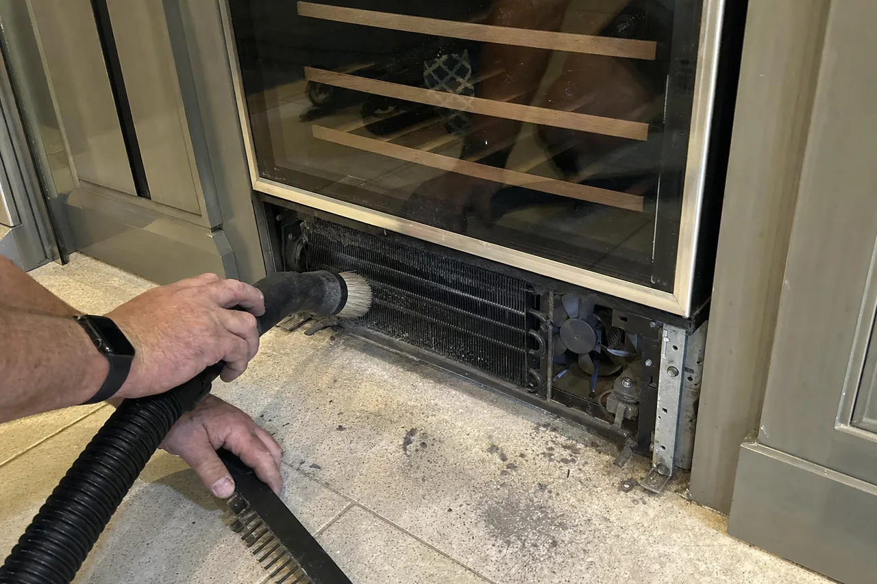 A technician drawing a vacuum brush along the condenser coil beneath a glass-door wine cabinet set into panelled joinery.