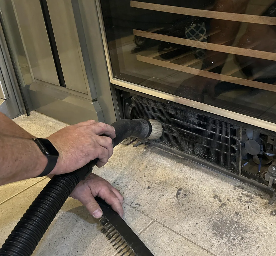 Hands guiding a brush nozzle across the condenser coil under a glass-fronted undercounter cabinet, dust visible on the tile in front of it.