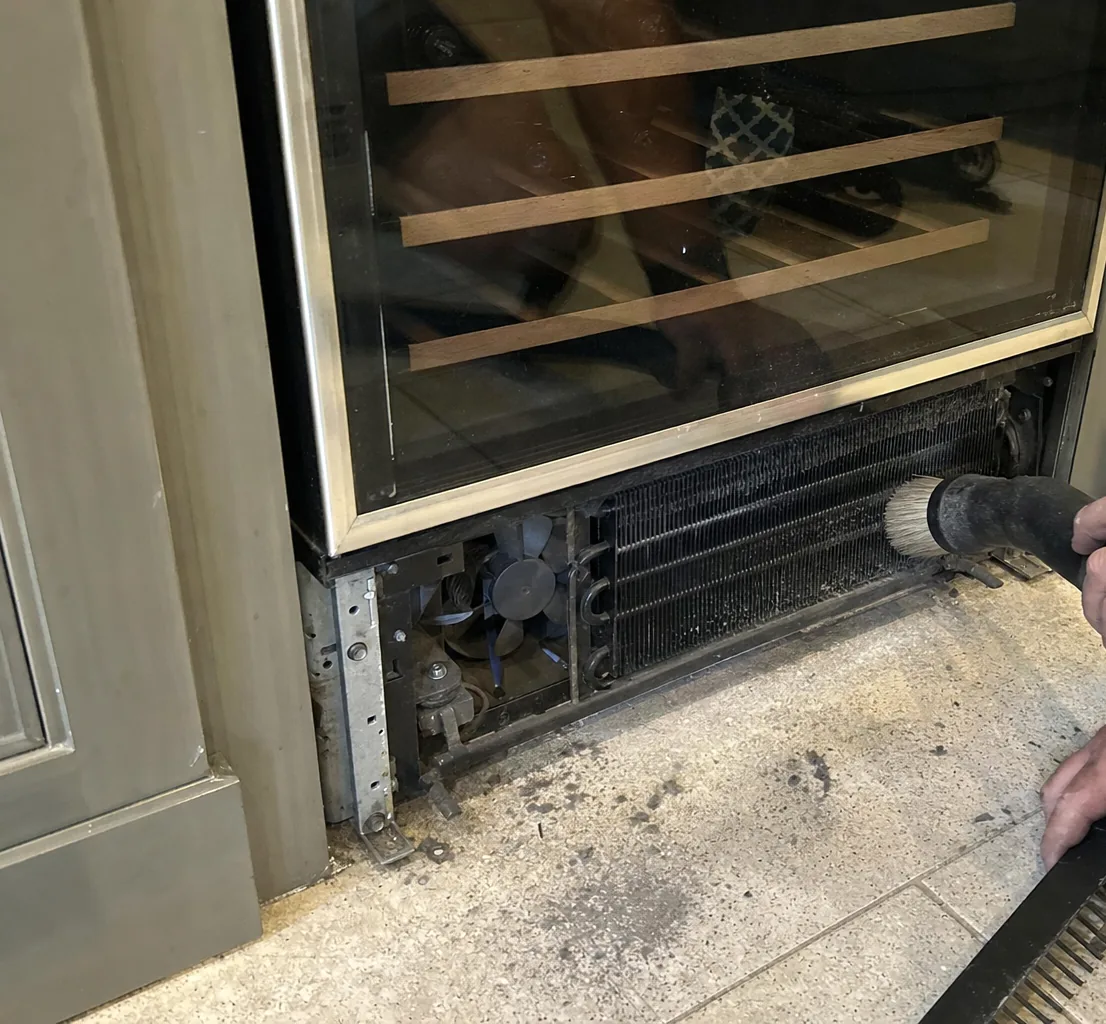 A glass-fronted wine cabinet with wooden shelf runners visible behind the door, its lower grille removed for a coil clean.