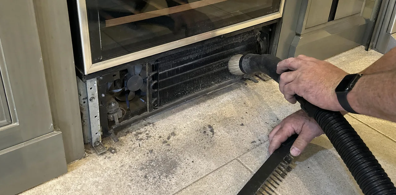 A wide, low view along the floor at an open toe-kick grille, condenser fins and fan exposed, brush nozzle in hand.
