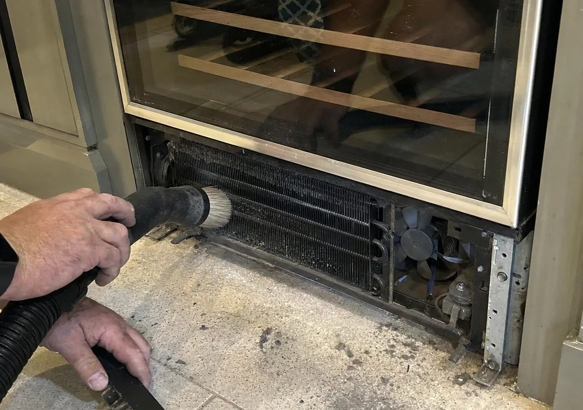 The base of an undercounter cabinet with its grille off, condenser coil and fan exposed a few inches above the tile.