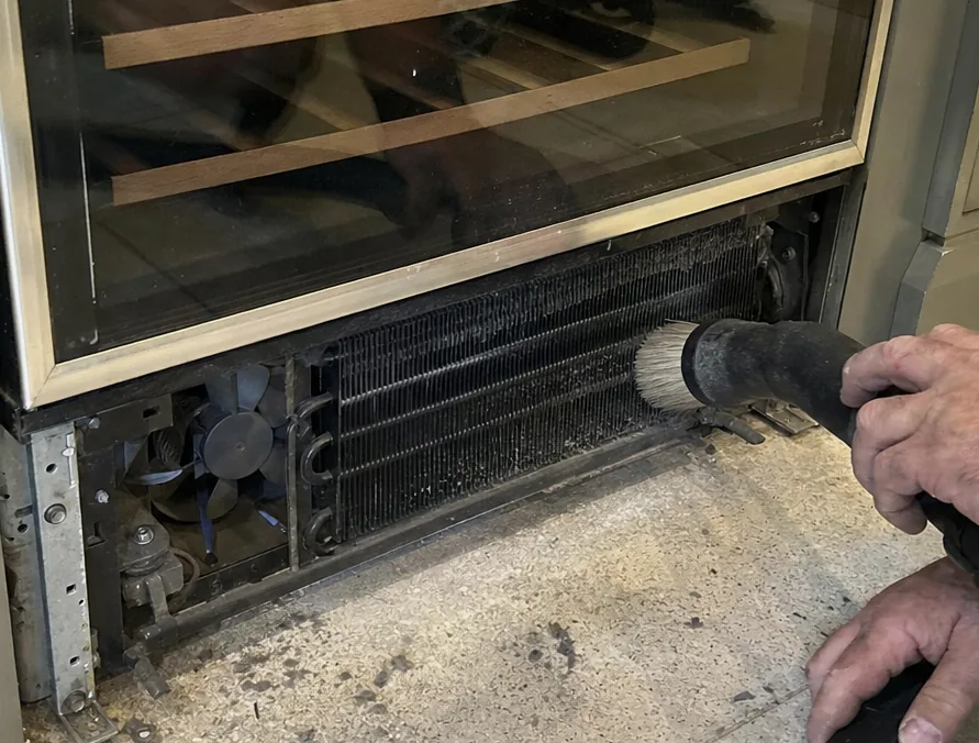 A tight view of a condenser coil and its fan behind an open toe-kick grille, a brush nozzle entering from the right.