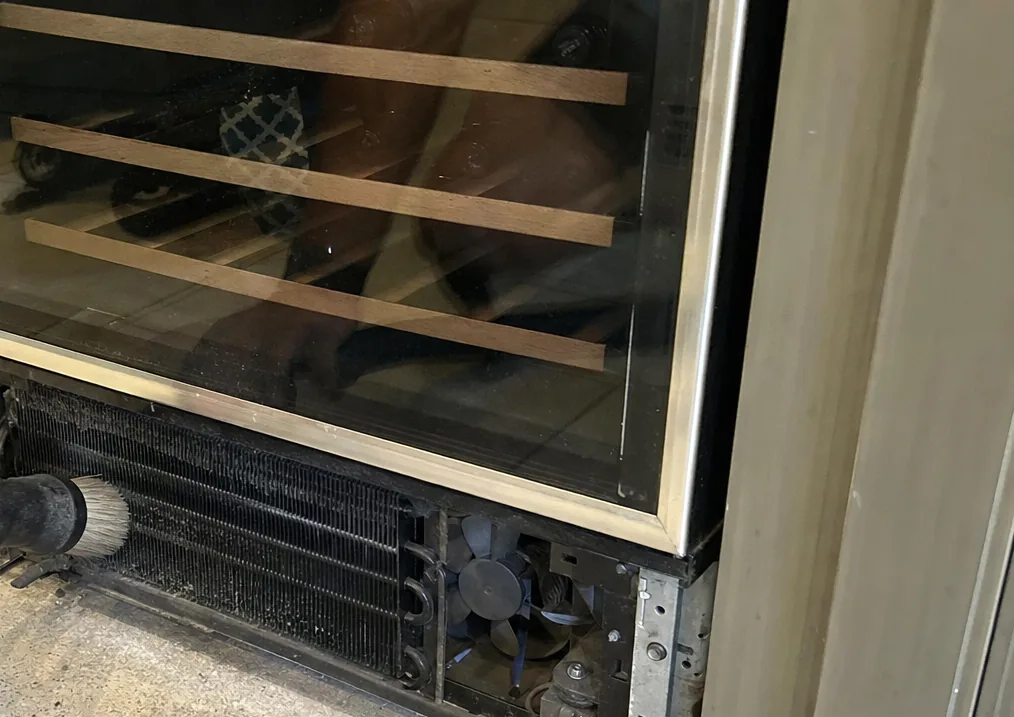 Bottles racked on wooden runners behind the smoked glass of a wine cabinet, its lower grille removed to show the coil.
