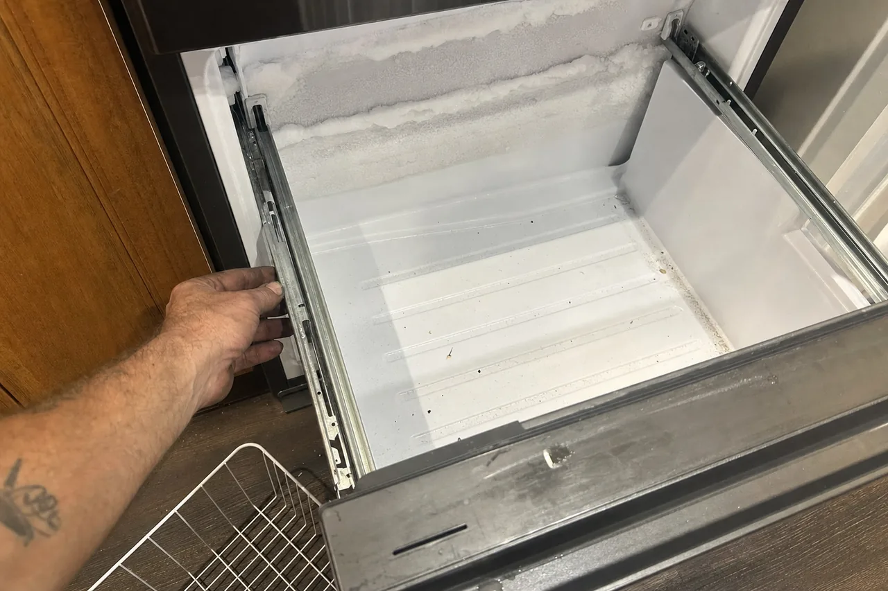 A hand pulling a freezer drawer open on its slides, frost along the rear wall and an empty wire basket on the floor beside it.