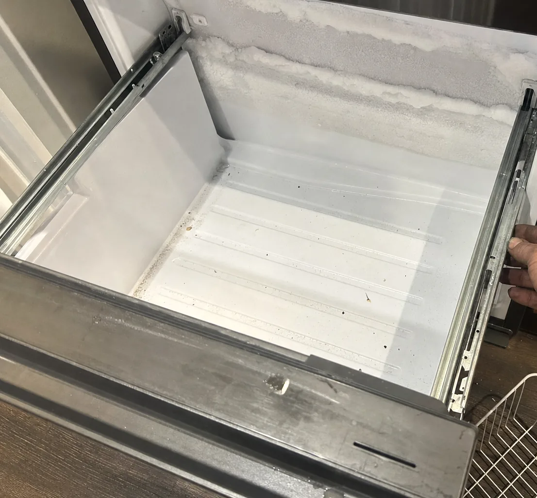 An open freezer drawer with a bare liner and a bank of frost along its back wall, the drawer front tilted forward.