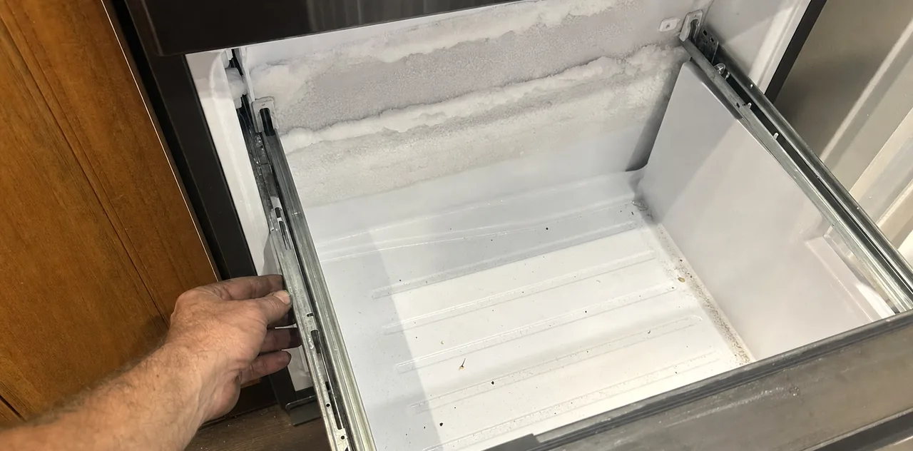 A wide view into an empty freezer drawer, frost banked across the back wall and a wire basket set out on the floor.