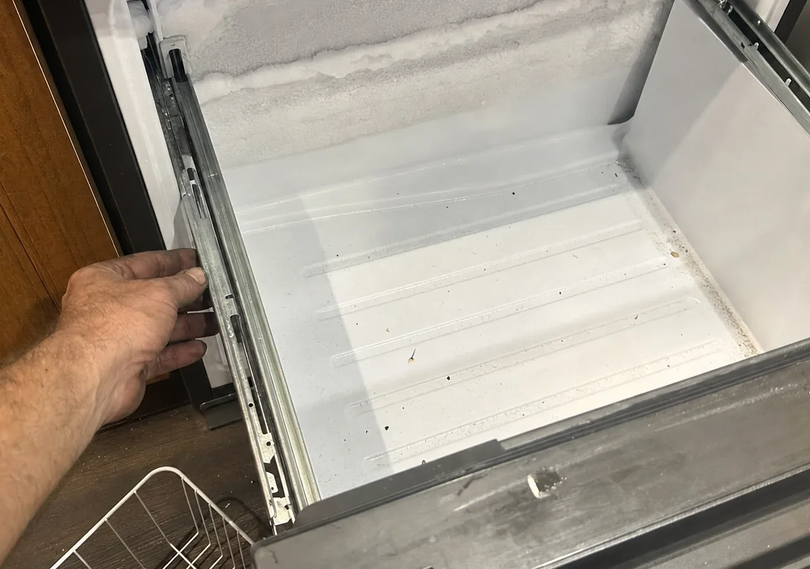 A hand on the rail of an open freezer drawer, the frost line along the compartment wall clearly visible above it.