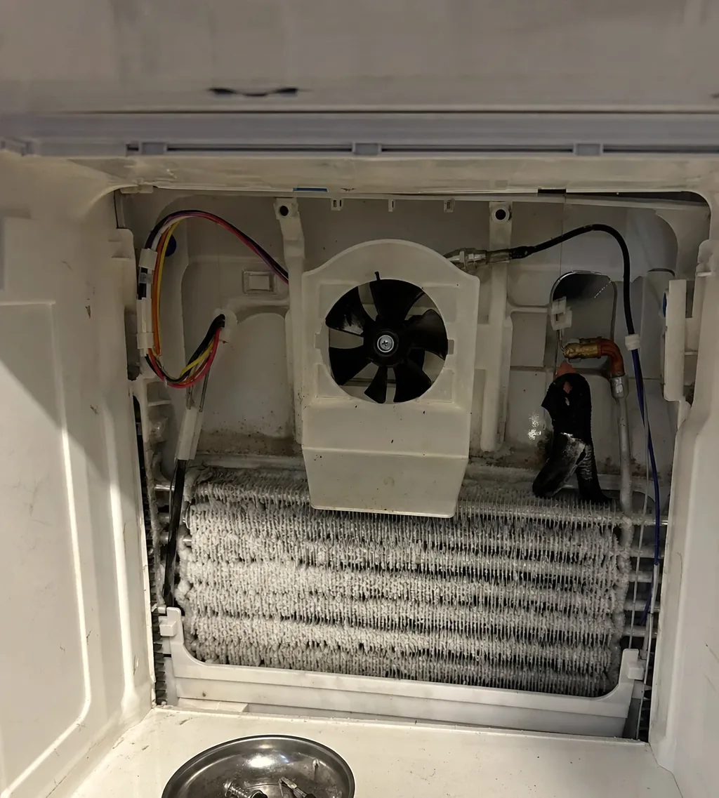 The rear panel of a freezer removed to show the evaporator coil packed with white frost and the fan housing above it.