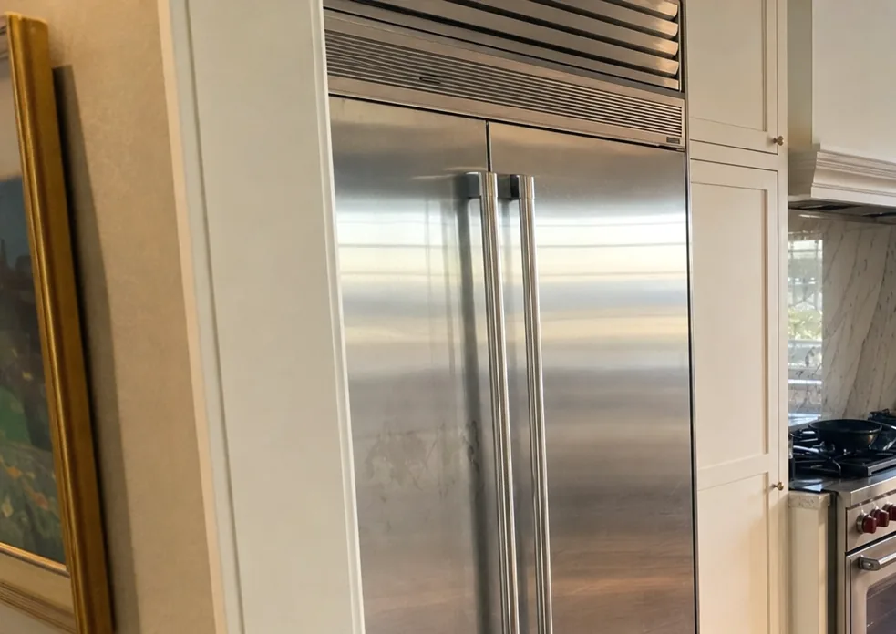 The upper grille and handles of a built-in refrigerator seen close, with a framed picture on the wall beside it.