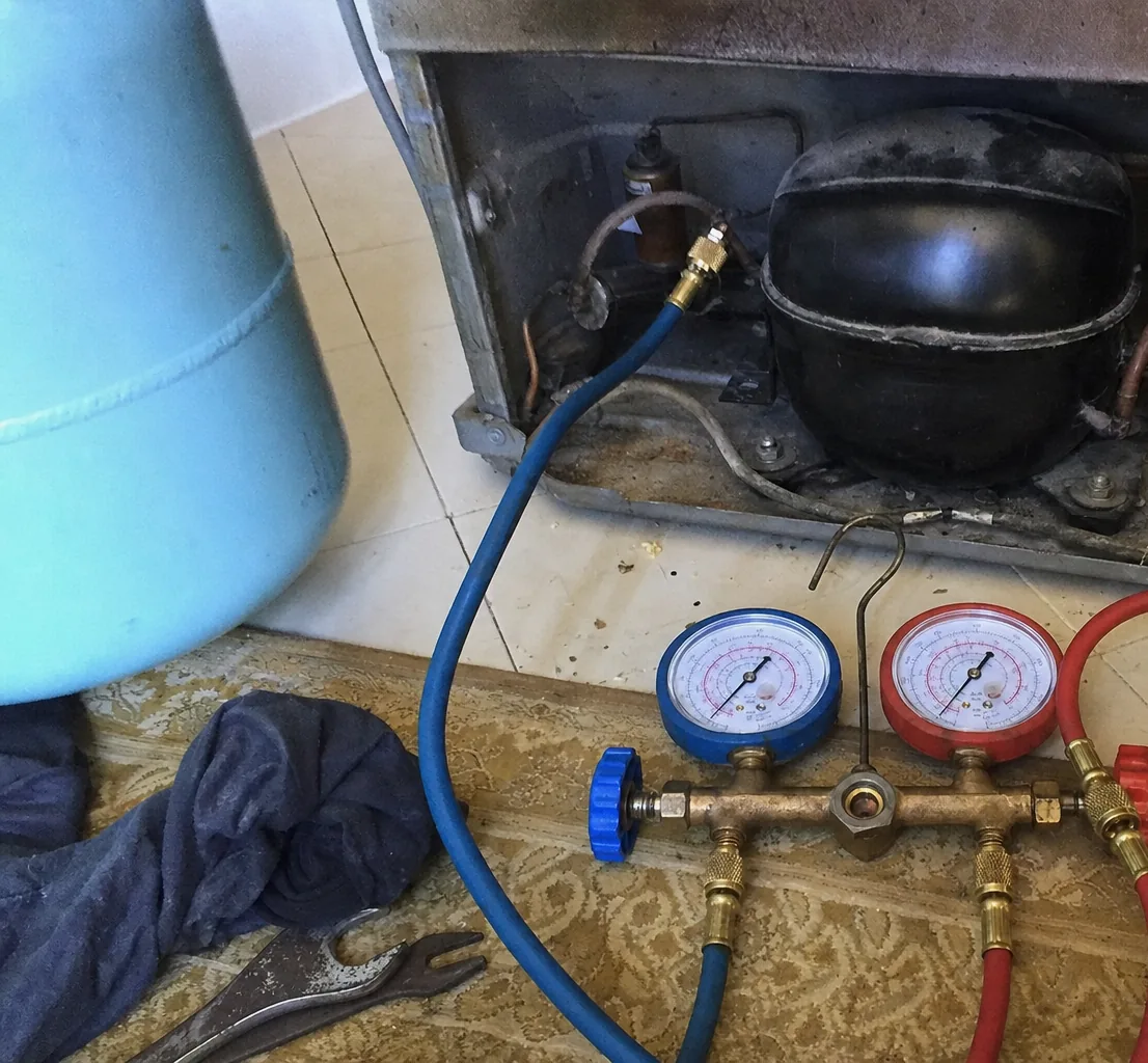 A refrigerant manifold with blue and red gauges standing on the floor, its hoses running up into the machine compartment of a cabinet.