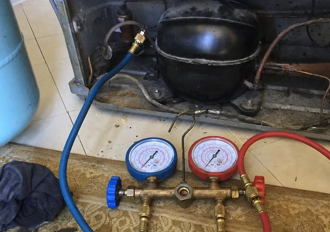 A close view of a compressor and its copper lines in the machine compartment, with the gauge manifold laid on the floor below.